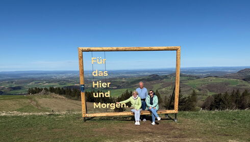 Drei Personen sitzen in Holzbilderrahmen vor Berglandschaft